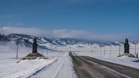 The highway goes off into the distance through a snow-covered valley. White markings on the asphalt. The figures of idols stand on the roadsides. Ahead, against the sky-picturesque mountains. Altai.の写真素材