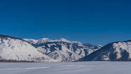 Pure white snow covers the valley. A lonely path is trampled through snowdrifts. A picturesque mountain range against a clear blue sky. Altaiの写真素材