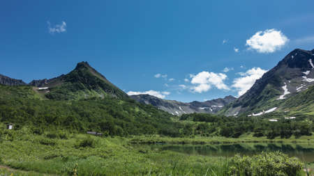 A calm lake on an alpine meadow. Lush green vegetation on the banks. A picturesque mountain range against a background of blue sky and clouds. Kamchatka. Vachkazhets. Lake Tahkolocの写真素材