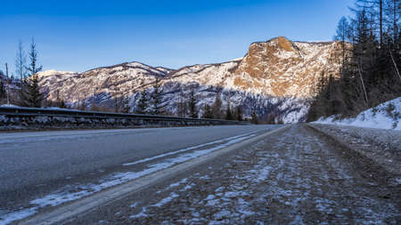 The highway goes forward, towards the sunlit mountains. Snow on steep slopes. blue sky. There is a...