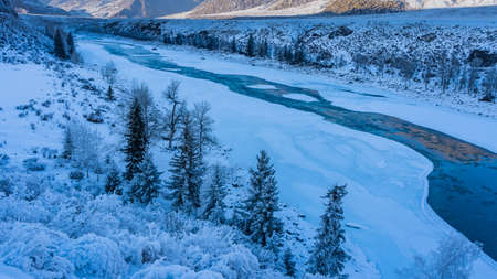 A blue ice-free river flows between the snow-covered shores. Coniferous trees and bushes are covered...