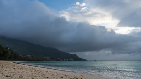 A foggy day on a tropical beach. The turquoise ocean is calm. Foam of waves on the sand. A rain cloud hung over the hill. Seychelles. Mahe. Beau Vallonの写真素材