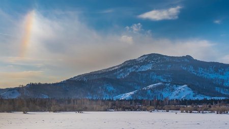 The forest grows in a snowy valley. A mountain range against a blue sky. An optical effect is visible - a band of rainbow glow in the clouds. Altaiの写真素材