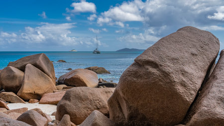 A yacht is visible in the calm turquoise ocean. The outline of the island on the horizon. In the foreground is a pile of granite boulders. Blue sky with clouds. Seychelles. Praslinの写真素材