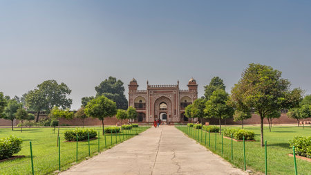 The entrance to the ancient tomb of Itmad-Ud-Daulah. The red sandstone walls are decorated with white ornaments. People walk along the path through the arched gate. Grass and treesの写真素材