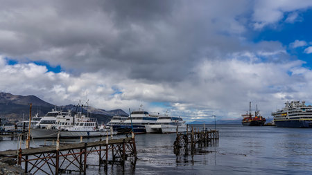 The seaport of Ushuaia. Cruise ships and pleasure boats are moored in the bay. Mountains against a background of blue sky and clouds. An old weathered pedestrian deckの写真素材