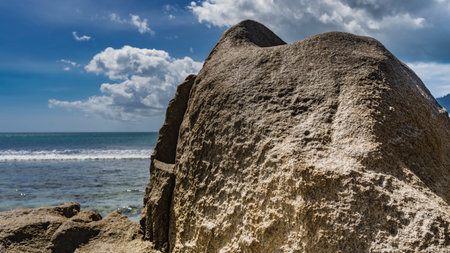 Granite rough rock with bizarre outlines rises on the shore of the turquoise ocean. The waves are foaming in the distance. Clouds in the blue sky. Seychelles. Mahe.の写真素材