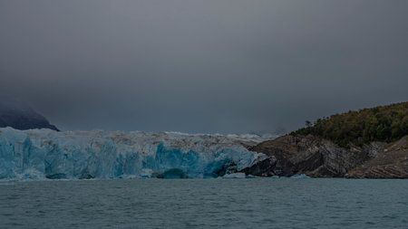 Perito Moreno Glacier. Blue ice over the turquoise water of the lake. There is green vegetation on the coastal rocks. cloudy. The mountains are hiding in the fog. Argentina.の写真素材