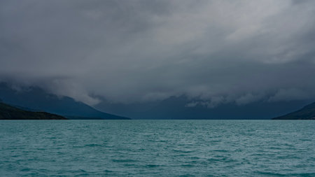 A beautiful turquoise glacial lake is surrounded by mountains. The peaks are hidden in fog and...
