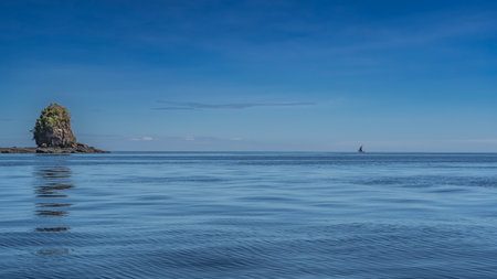 A picturesque rocky island with steep slopes is visible in the ocean. Trees grow on the top. Reflection in the water. In the distance, against the blue sky, a sailing boatの写真素材