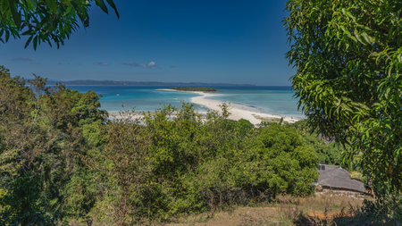 Beautiful postcard view of a tropical island covered with green vegetation. A winding sand spit in the turquoise ocean. Boats in the water. In the foreground are tree branches,の写真素材