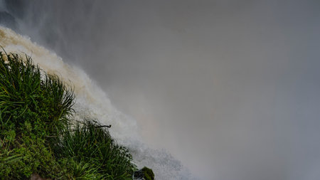 A stormy stream of water falls into the abyss from the cliff. The waterfall foams, splashes in the air. Everything is shrouded in thick fog. In the foreground is lush green grass.の写真素材