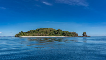 The island in the ocean is covered with green vegetation. Nearby is a picturesque rock. Tiny silhouettes of people, moored boats on the sandy beach. Blue shiny water, azure sky.の写真素材