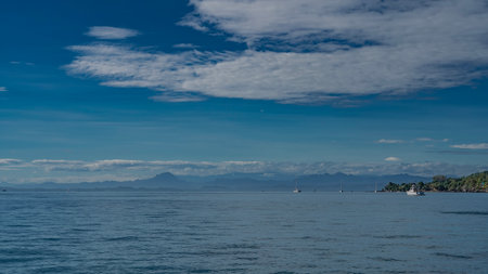 Seascape. Boats are visible in the calm blue ocean. Green vegetation on the shore. A mountain range in the distance. Azure sky with white clouds. Madagascar. Nosy Kombaの写真素材