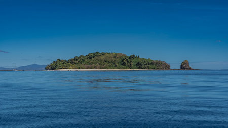 A picturesque tropical island and a rock against the background of blue sky and ocean. Lush green vegetation on the hill. Boats, tiny silhouettes of people are visible on the beachの写真素材