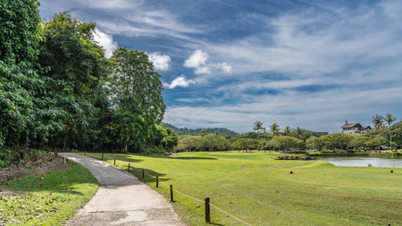 A concrete footpath winds along the golf course. Rope railings along the path. Golf balls are laid out on the green grass of the lawn. Lush tropical vegetation. The lakeの写真素材