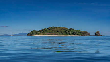 The beautiful tropical island is completely covered with lush green vegetation. The rock is nearby. Ripples on the brilliant blue ocean water. The yacht is far away. Mountainsの写真素材