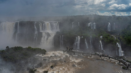 A pedestrian bridge and a tourist observation deck are laid over the stormy river. Cascades of waterfalls descend from the cliffs into the riverbed. Splashes, fog. Cloudsの写真素材