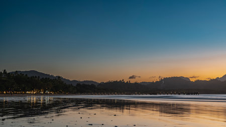 Twilight after sunset in the tropics. Silhouettes of mountains against the blue-orange evening sky. Lights of lanterns in the distance. Ocean waves are spreading over the beach.の写真素材