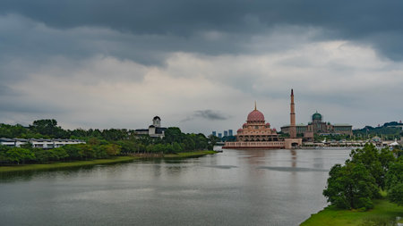 A calm lake in the city park. Green vegetation on the banks. A beautiful pink mosque is visible in the distance. Domes and minaret against a cloudy sky. Malaysia. Putrajaya. Putraの写真素材