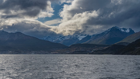 The beautiful Andean Martial mountain range. Snow-capped peaks against the sky and clouds. The city buildings of Ushuaia are visible on the ocean. Ripples on the water of theの写真素材