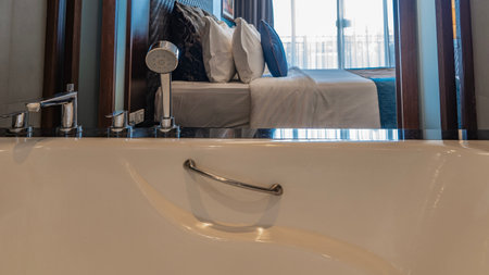 There is a stainless steel faucet, shower and handrails on the edge of the white acrylic bathtub. The bedroom is visible through the open door. The bed is covered with sheets.の写真素材