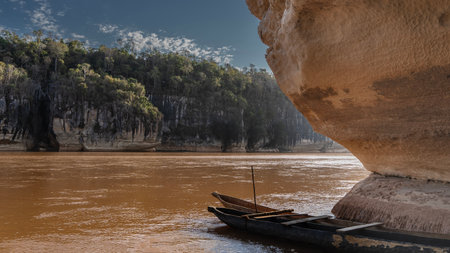 Canoes hollowed out of wood are moored at the entrance to the cave. The rock overhangs the red-brown river. Green vegetation on the far steep bank. Blue sky, clouds. Madagascar.の写真素材