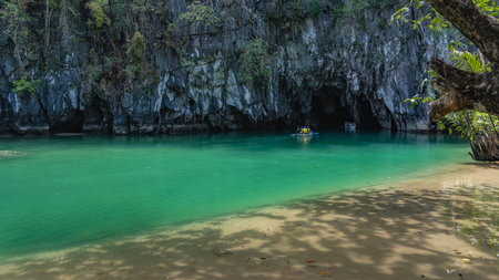 A sheer cliff above a calm turquoise river. Canoes with tourists sail towards the dark entrance of the cave. Philippines. Palawan. Puerto Princesa Subterranean River National Parkの写真素材