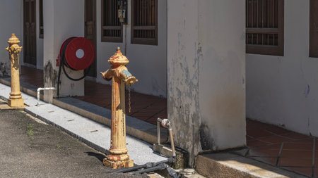 Old fire hydrants on the road outside the building. The yellow paint is peeling off. The rusty chain is hanging down. The hose is rolled up, suspended from a worn wall. Malaysia.の写真素材