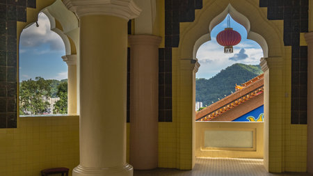 A red Chinese lantern is suspended in the elegant arched vault of the temple window opening. Through the hole, you can see a fragment of the roof with ornaments, blue sky, clouds,の写真素材