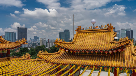 Bright tiled roofs of a Chinese temple against the city skyscrapers, blue sky, clouds. Traditional red and yellow lanterns are suspended along the curved edges.の写真素材