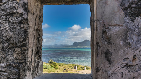 Through the window opening, you can see the turquoise ocean, mountains, blue sky, clouds, a hillside with green grass. The landscape is surrounded by a frame of weathered, crackedの写真素材