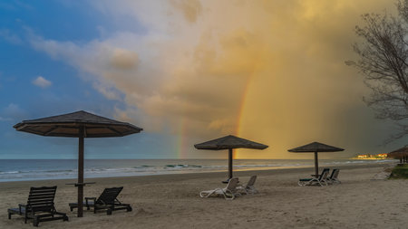 Beautiful double rainbow over the ocean. The blue sky and clouds are illuminated by golden evening light. A row of sun loungers on a sandy beach under an umbrellas. The wavesの写真素材