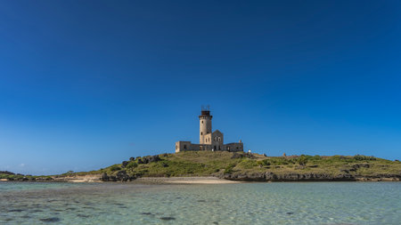 The old lighthouse on the island. A dilapidated building with a tall tower against a clear blue sky. Transparent turquoise ocean in the foreground. Copy Space. Mauritius.の写真素材