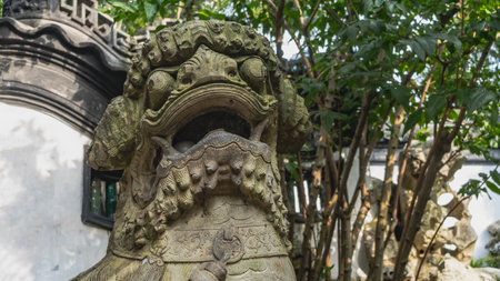 Sculpture of a mythical lion. A fragment of a statue. A head with an open mouth and large fangs. Carved decorative ornaments. China. The background is green vegetation. Front view.の写真素材