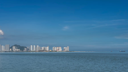 View of the city embankment from the ocean. Tall multi-storey buildings, skyscrapers, a green hill against the azure sky and clouds. Ripples on the surface of the blue sea.の写真素材