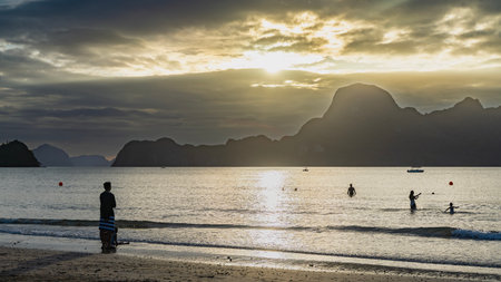 A serene sunset in golden tones. A tropical island. The sun is shining from behind the clouds. Mountains against the sky. Silhouettes of people in the ocean. A sunny pathの写真素材