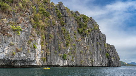 A tourist canoe floats in the ocean along a sheer cliff. Green vegetation on steep furrowed slopes....