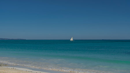 A lonely boat is floating in a calm turquoise ocean. A white sail against a clear blue sky. Foam...