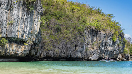 A traditional double-outrigger dugout Filipino bangka boat is anchored in the turquoise ocean next to the steep coastal cliffs. Green tropical vegetation on the slopes.の写真素材