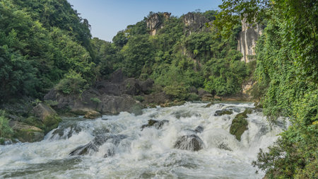 A stormy mountain river. Rushing streams of water foam on boulders and rapids. Lush green vegetation on the shores. Rocks against the blue sky. China. Tianxingqiao Scenic Area.の写真素材