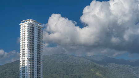 A white skyscraper against a background of blue sky, clouds, and a green hill. A tall multi-storey tower. Windows, balconies on the facade. Roof terrace. Malaysia. Penang.の写真素材