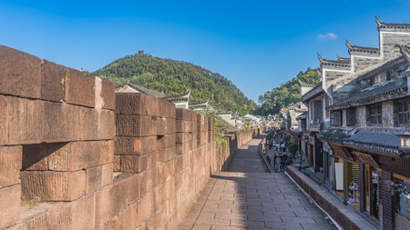 The ancient brick fortress wall of Fenghuang Old Town. Traditional Chinese buildings along a paved street. People are walking on the sidewalk. The mountain against the blue sky.の写真素材