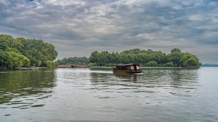 A beautiful Chinese boat is sailing on the lake. There is a wooden pavilion with a tiled roof on the deck. Green vegetation on the shore. Stone arch bridge in the distance.の写真素材