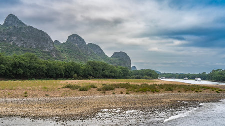 A picturesque river. On the pebbly shores there is green grass, foam of waves and trees. Tourist sail ships in the distance. Mountains against the sky and clouds. China. Lee River.の写真素材