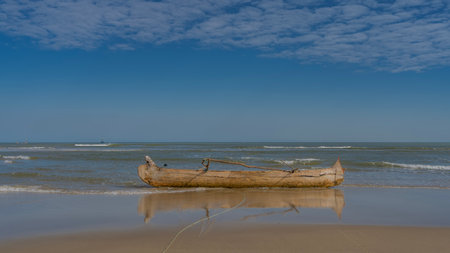 Low tide in the ocean. An old unpainted pirogue boat in shallow water. A crack on the side. Reflection on wet, smooth sand. Turquoise sea waves are foaming. Blue sky, clouds.の写真素材