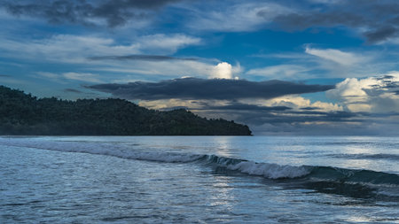 Morning seascape. Twilight. The ocean waves roll towards the shore, foam and spread. The clouds in the blue sky are illuminated with golden light. A hill in the distance. Malaysiaの写真素材