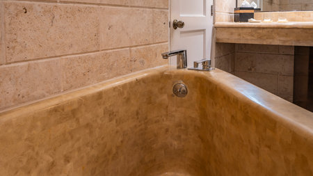 A fragment of the bathroom. A triangular beige bathtub in the corner against a tiled wall. Shiny metal faucets on the edge. Glasses, napkins, soap on the countertop. The doorの写真素材