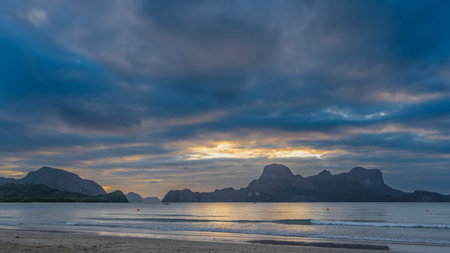 Beautiful tropical sunset over the ocean. The blue clouds in the sky are highlighted in pink. Golden glow. Waves are foaming on a sandy beach. Silhouettes of mountainsの写真素材