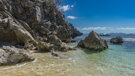 Tropical seascape. Foam on the clear turquoise ocean water. Picturesque boulders and steep coastal cliffs. The silhouette of a bangka boat in the distance. Blue sky, clouds.の写真素材